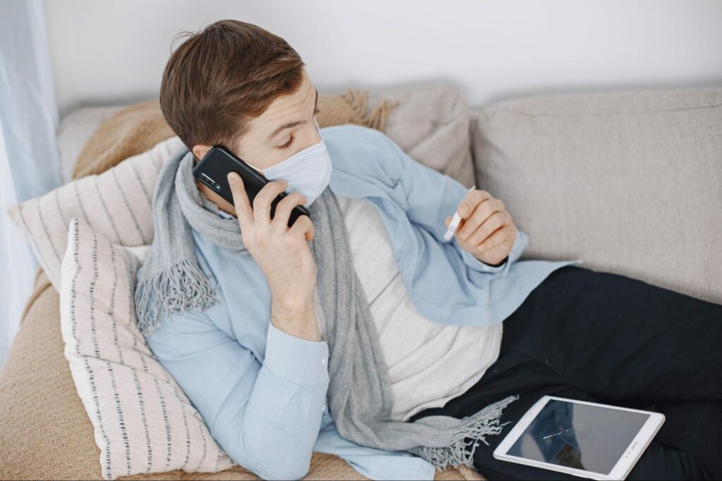 Man resting on a couch with a thermometer and mask while calling an after hours clinic phone number for medical advice