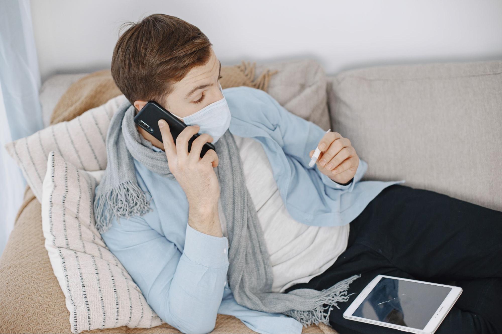 Man resting on a couch with a thermometer and mask while calling an after hours clinic phone number for medical advice