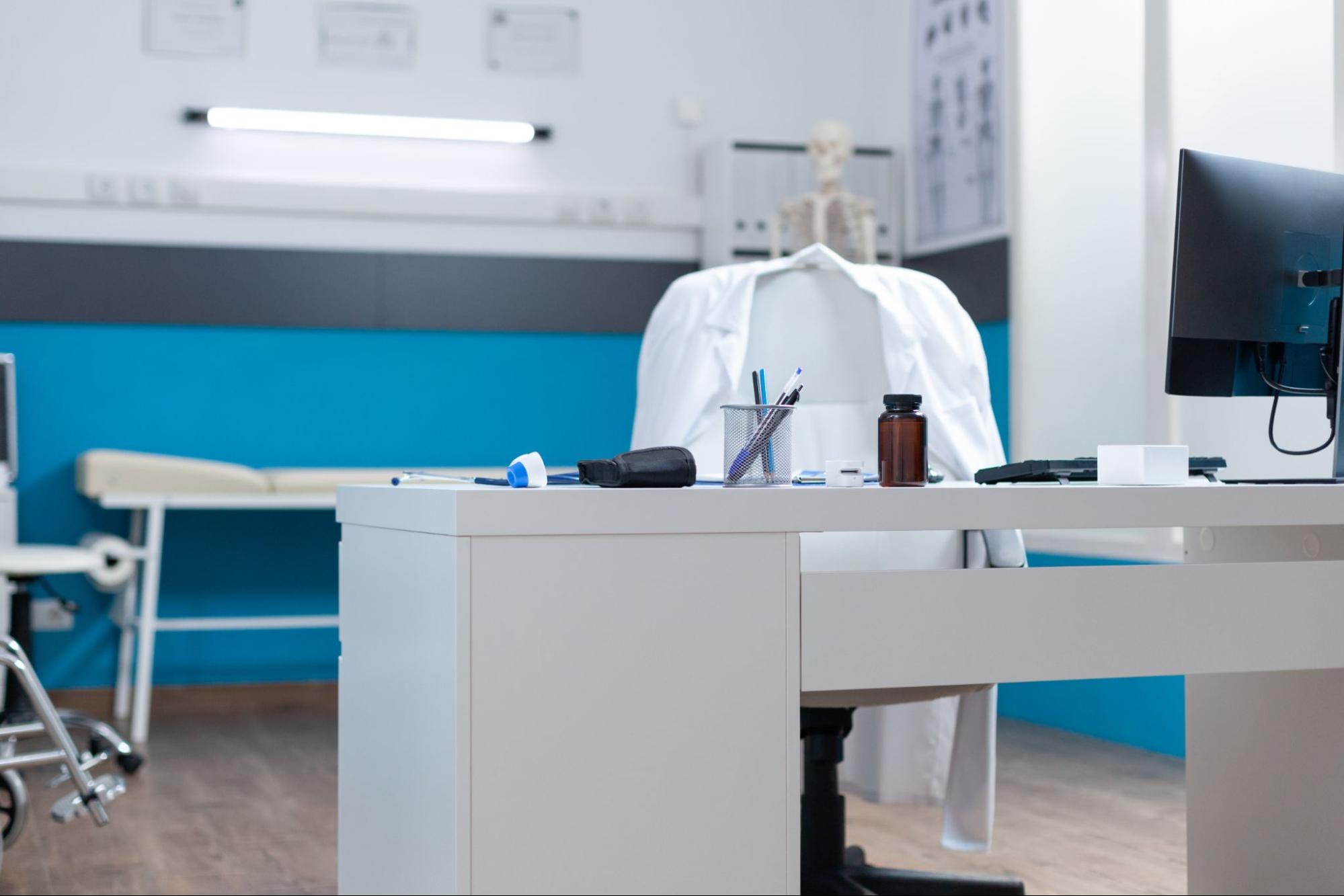 Empty medical exam room with desk, equipment, and exam table 