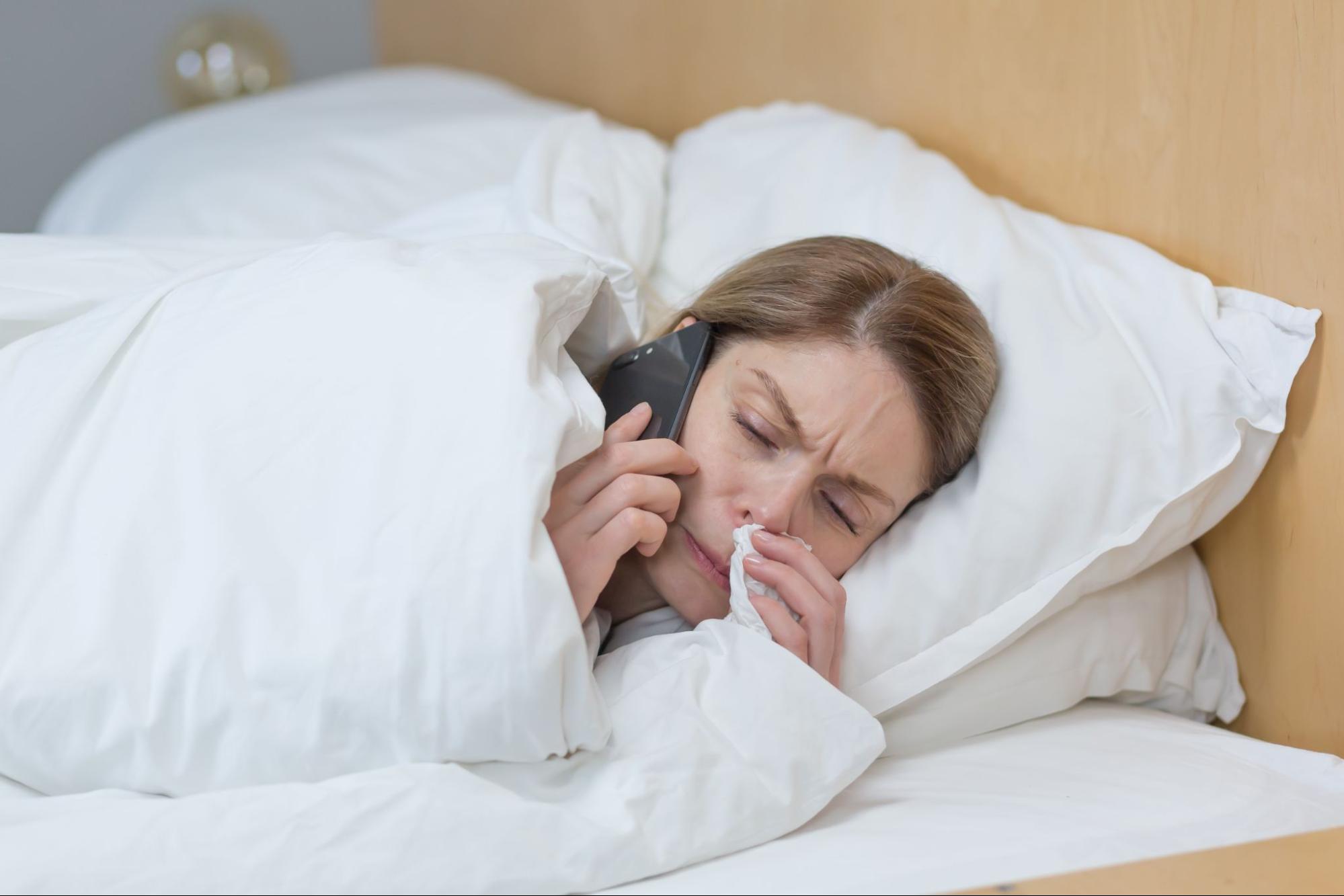 Sick woman in bed with tissue calling an after hours clinic phone number for medical guidance
