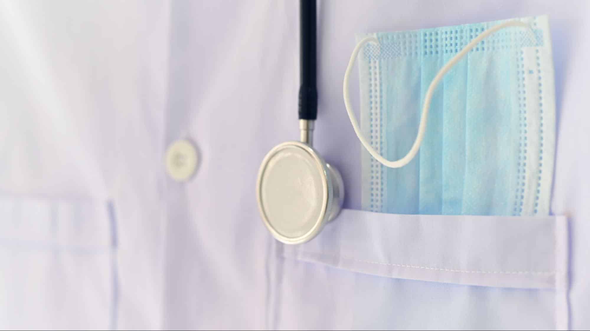 Close-up of a white lab coat pocket containing a blue surgical mask and a stethoscope hanging nearby, representing primary care near me in weslaco tx