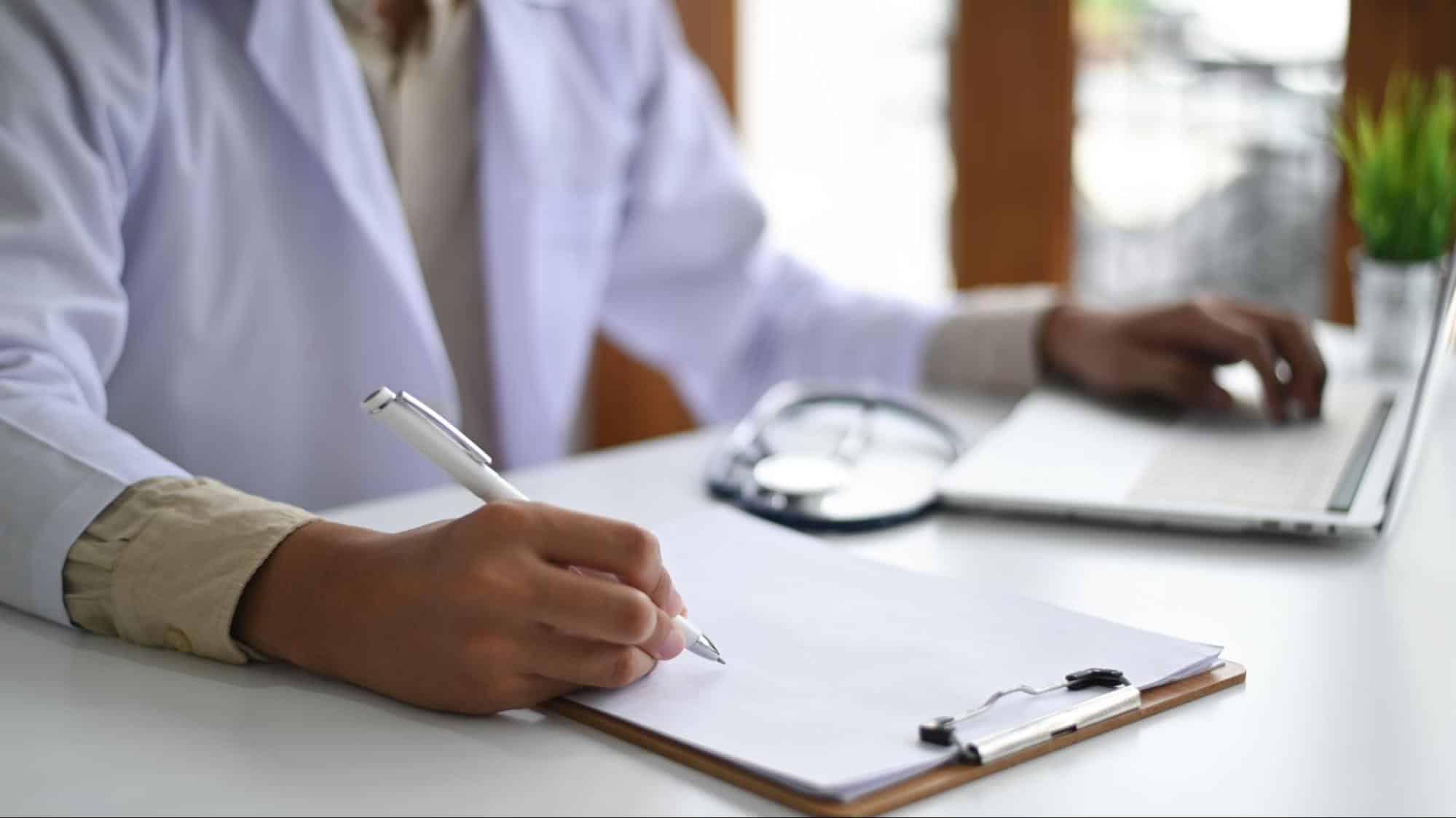 A healthcare professional in a white coat writing on a clipboard while working at a laptop.